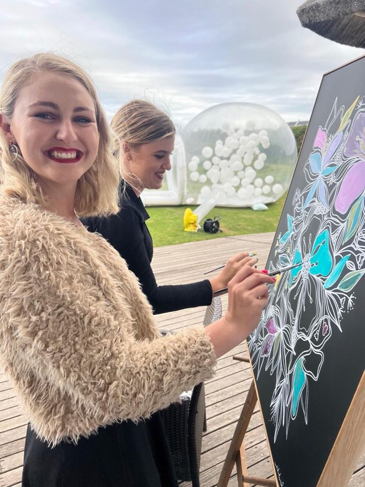 Two guests smiling while they paint a floral artwork at an outdoor event