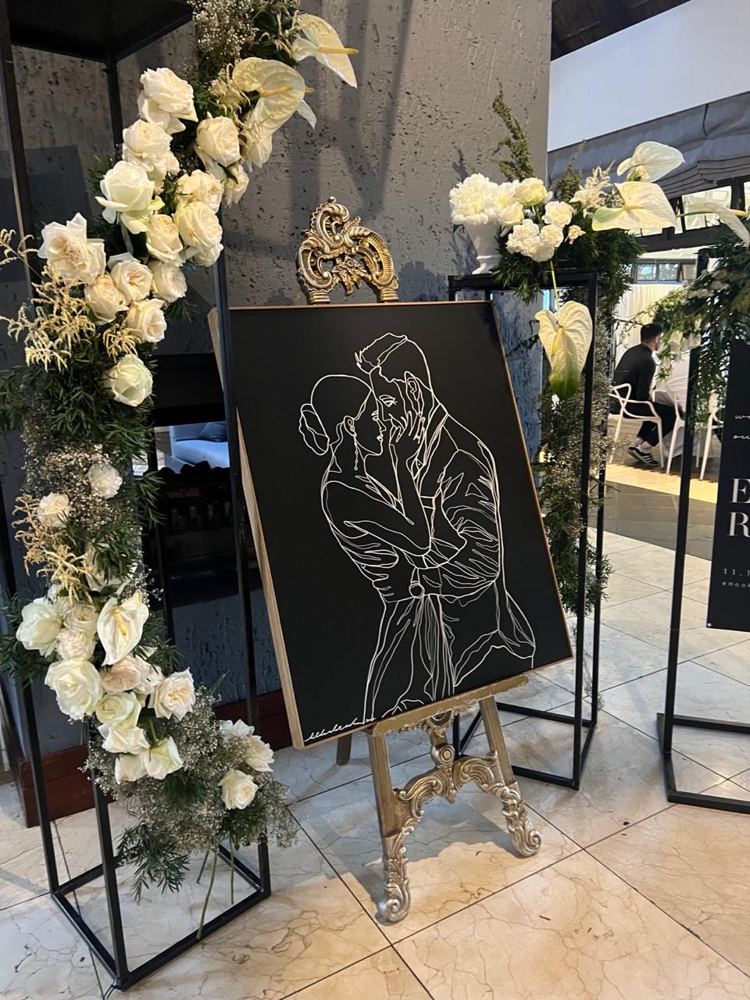 A line portrait of a bride and groom on a gilded easel, framed by white roses at a wedding